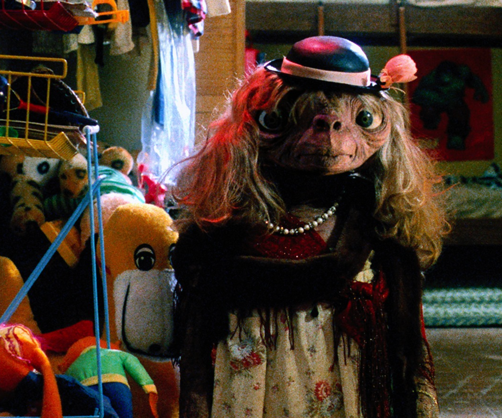 Still from a scent of the 1982 movie ET, showing the eponymous ET wearing a wig, dress, bowler hat, shawl, jewelry